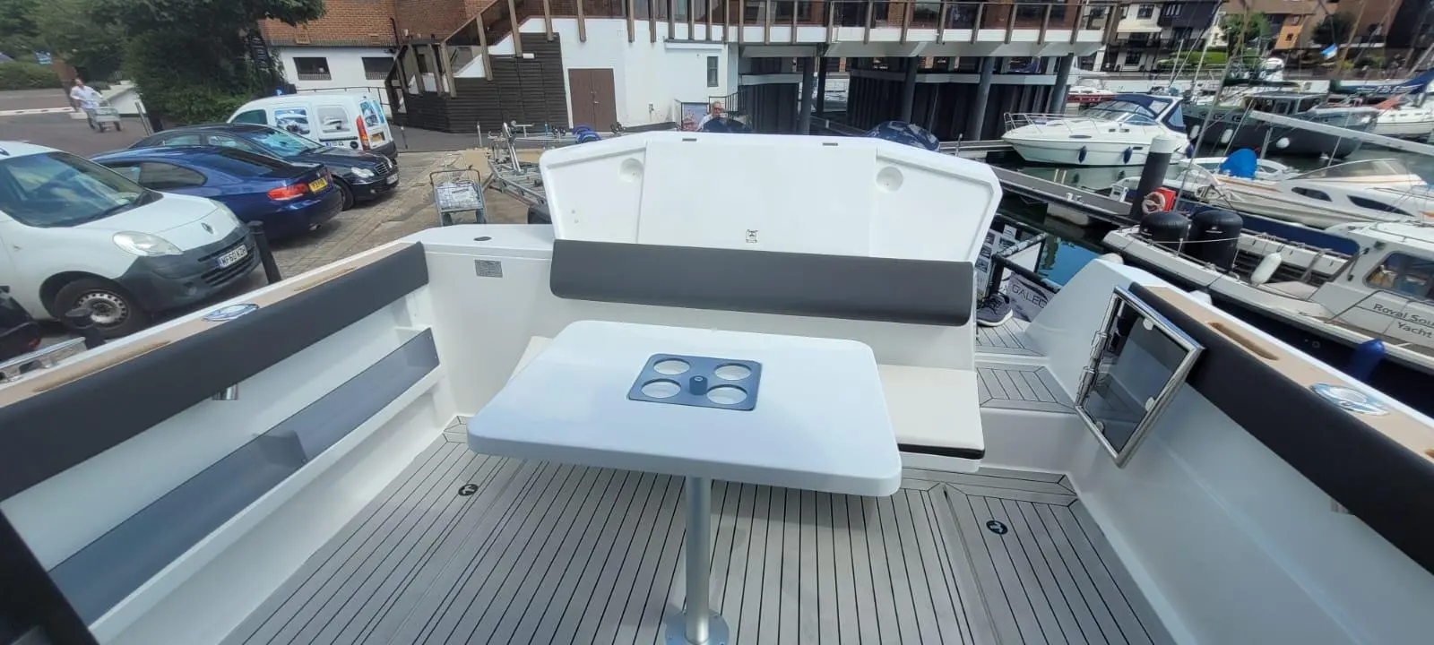 Interior of a boat featuring a white table with drink holders, a grey cushioned bench, and a marina view.